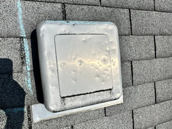 Metal roof vent with visible surface dents surrounded by shingles